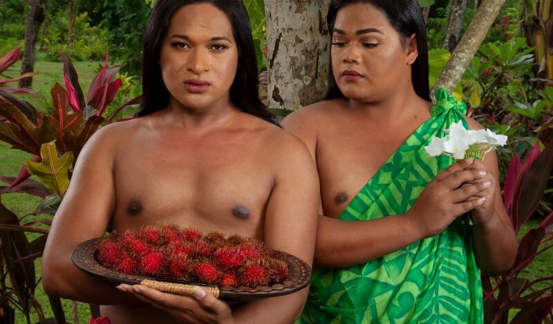 Two people stood next to one another wearing traditional Pacific dress. One person wears a red sarong and holds a plate of red fruit and the other wears a green dress and holds a white flower.