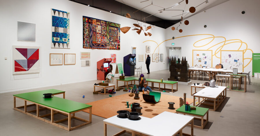 Green and white benches surround an orange play area. Along the walls are Minecraft Machines, and various artworks from the Whitworth's collection.