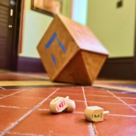 A wooden spinning top with four sides with a different Hebrew letter on each side, also known as a driedel, on a tiled floor.