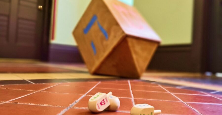 A wooden spinning top with four sides with a different Hebrew letter on each side, also known as a driedel, on a tiled floor.