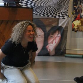 A young person kneeling on the floor with her hands behind her back, leaning forward and laughing like she is really happy.