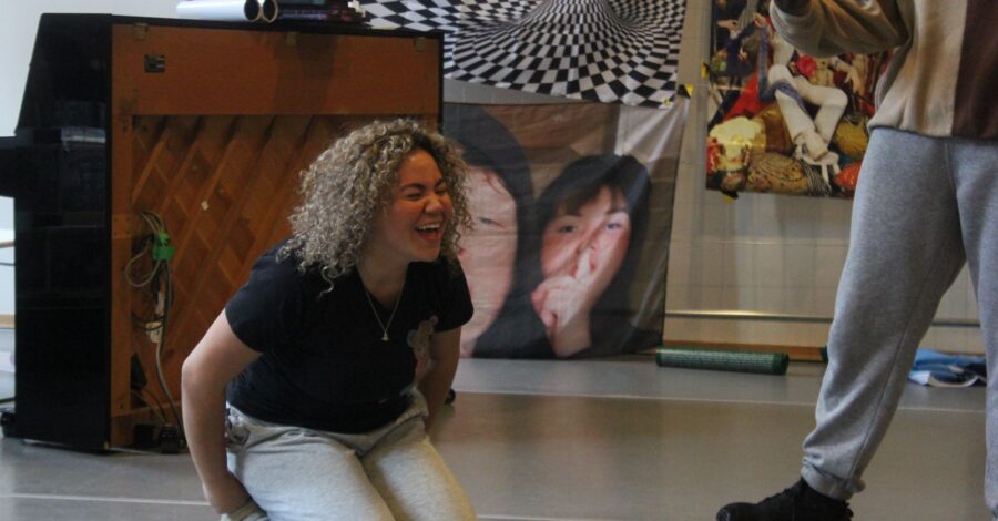 A young person kneeling on the floor with her hands behind her back, leaning forward and laughing like she is really happy.