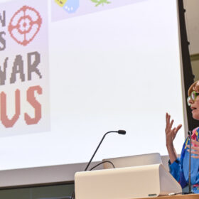 A speaker, Dr Lisa Williams, talking at the National Chaordic Symposium about Portraits of Recovery. She stands in front of a large white screen by a desk with a microphone.