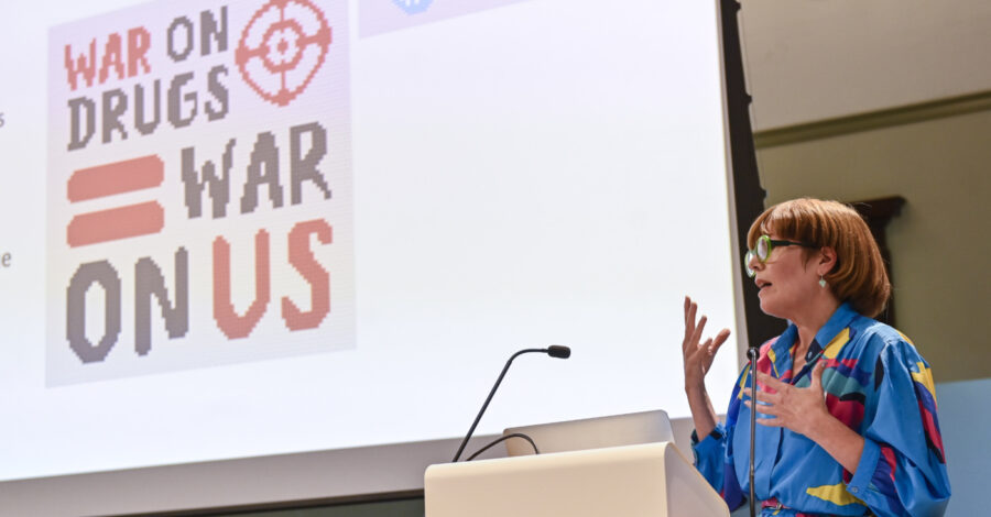 A speaker, Dr Lisa Williams, talking at the National Chaordic Symposium about Portraits of Recovery. She stands in front of a large white screen by a desk with a microphone.