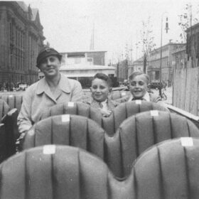 A black and white photograph showing a woman and two young boys are sitting on the top deck of a bus. They are smiling at the camera. The top deck is open and behind them we can see the streets of Berlin in the 1930s.