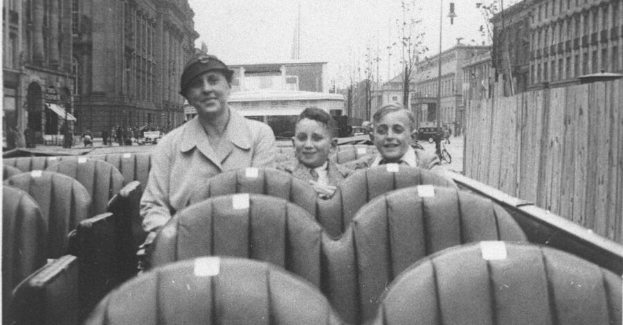 A black and white photograph showing a woman and two young boys are sitting on the top deck of a bus. They are smiling at the camera. The top deck is open and behind them we can see the streets of Berlin in the 1930s.