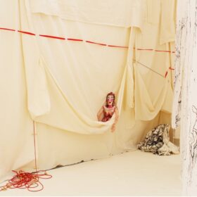 A doll with makeup peeks out of a hanging wall of butter yellow fabric. Red and black threads descend and cascade around the doll.