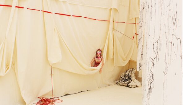 A doll with makeup peeks out of a hanging wall of butter yellow fabric. Red and black threads descend and cascade around the doll.