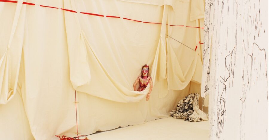 A doll with makeup peeks out of a hanging wall of butter yellow fabric. Red and black threads descend and cascade around the doll.