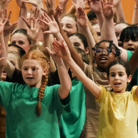 A choir of children wearing green and yellow t-shirts all raising their hands up into the air.