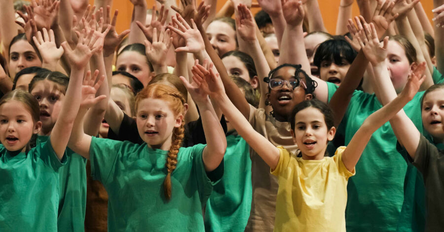 A choir of children wearing green and yellow t-shirts all raising their hands up into the air.