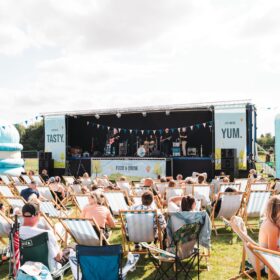 Lot of people sat outside on deckchairs. They all face towards a large outdoor stage.