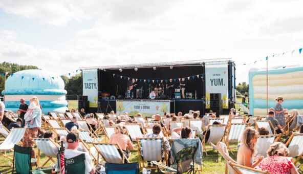 Lot of people sat outside on deckchairs. They all face towards a large outdoor stage.