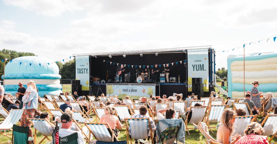 Lot of people sat outside on deckchairs. They all face towards a large outdoor stage.