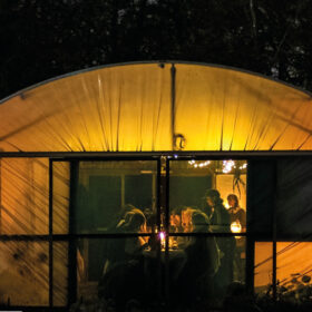 An tent lit up with warm lighting at night. There are people sat inside around a table.