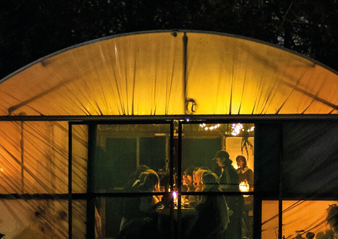 An tent lit up with warm lighting at night. There are people sat inside around a table.