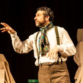 An actor with dark curly hair and a beard is acting on stage. He is wearing a white shirt, beige trousers with suspenders and a colourful scarf.
