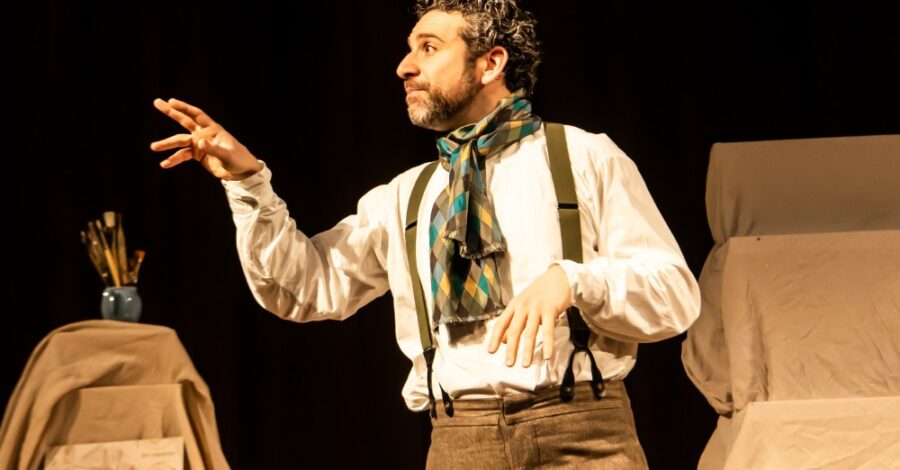 An actor with dark curly hair and a beard is acting on stage. He is wearing a white shirt, beige trousers with suspenders and a colourful scarf.