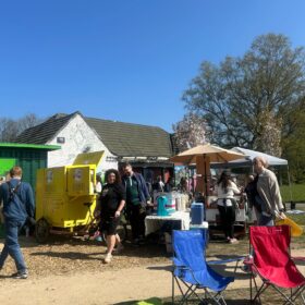 A small outdoor market with a few people bustling about. There are some parasols and also some chairs.