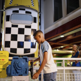The Experiment gallery at the Science and Industry Museum. A young person stands by a car that has been positioned vertcially. The car is yellow with a black and white chequered roof.