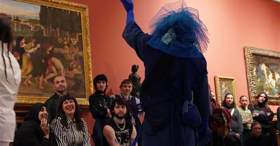 A performance in the gallery space. There is a performer standing with their back to the camera. They are dressed in black and have blue hair. Behind them, we can see the audience sat on chairs and laughing.