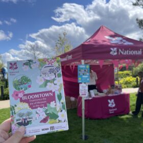 A hand holds a Bloomtown map in front of a pink National Trust gazebo on a sunny day