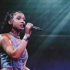 A singer performs on stage under dramatic lighting, holding a microphone close to her mouth. She wears a sleeveless top with layered jewellery, and beams of light shine down from above, highlighting her as she sings.