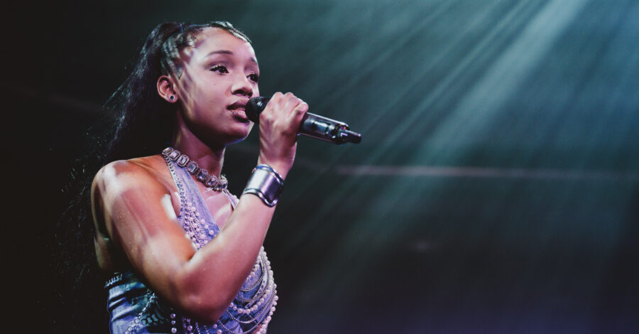 A singer performs on stage under dramatic lighting, holding a microphone close to her mouth. She wears a sleeveless top with layered jewellery, and beams of light shine down from above, highlighting her as she sings.