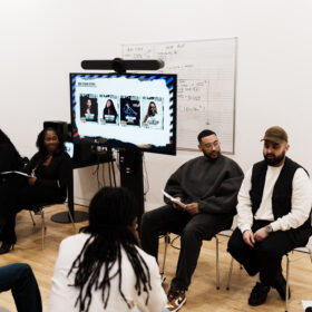 A group of people sit in a circle in a bright studio space during a discussion or workshop. A screen behind them displays event posters, while participants hold papers and listen to a speaker seated at the front.