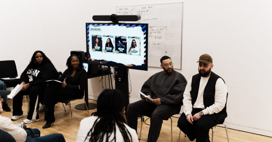A group of people sit in a circle in a bright studio space during a discussion or workshop. A screen behind them displays event posters, while participants hold papers and listen to a speaker seated at the front.