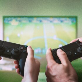 Two controllers and a two pairs of hands holding the consoles, with a screen in the background.