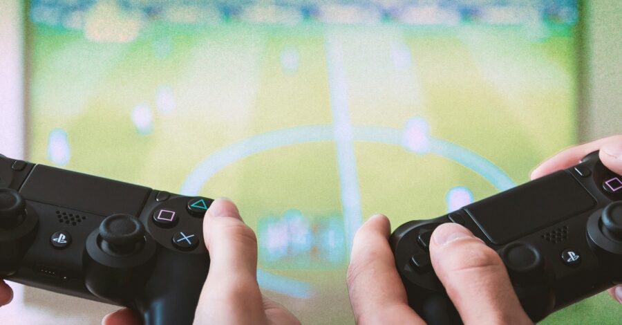 Two controllers and a two pairs of hands holding the consoles, with a screen in the background.