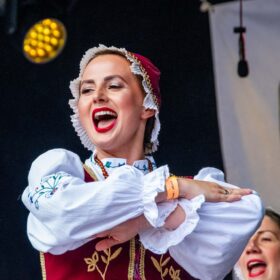 A woman wearing a colourful folk dance costume smiles as she performs a crossed arms pose on stage.