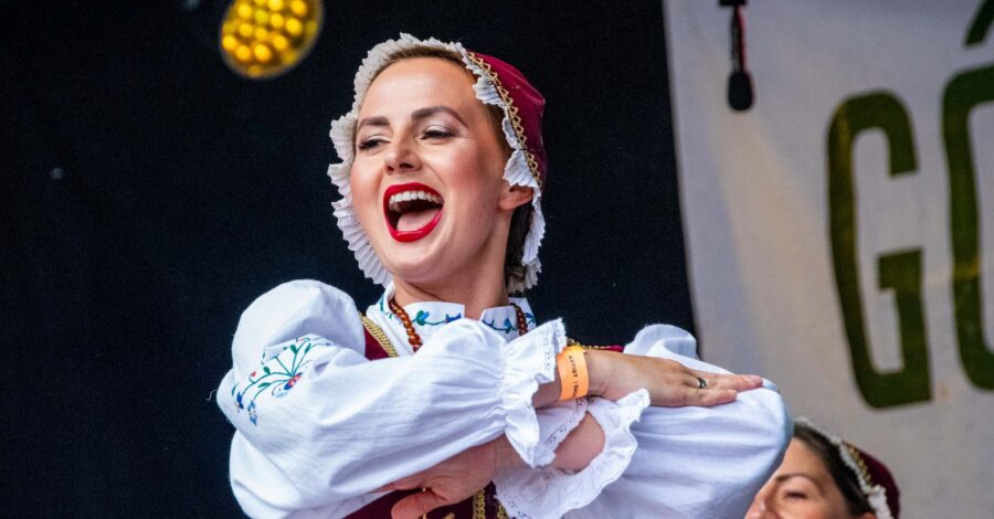 A woman wearing a colourful folk dance costume smiles as she performs a crossed arms pose on stage.