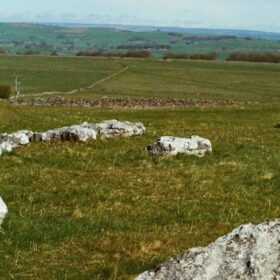 A wide countryside view shows pale limestone rocks scattered across a grassy slope, suggesting an ancient stone setting. A single person crouches beside one group of stones, while patchwork fields and low hills extend into the distance under a soft, open sky.