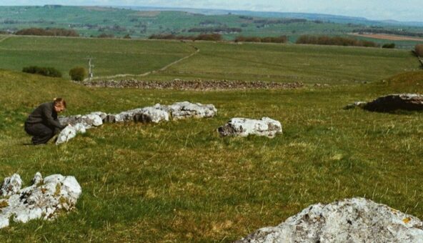 A wide countryside view shows pale limestone rocks scattered across a grassy slope, suggesting an ancient stone setting. A single person crouches beside one group of stones, while patchwork fields and low hills extend into the distance under a soft, open sky.