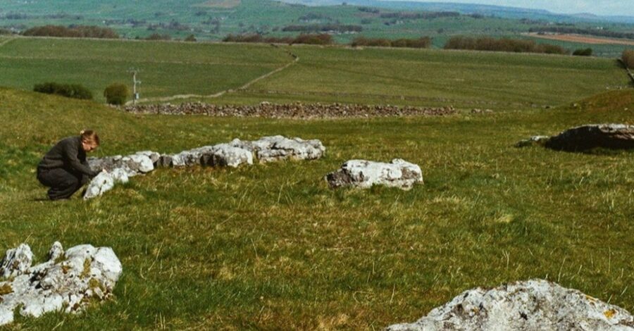 A wide countryside view shows pale limestone rocks scattered across a grassy slope, suggesting an ancient stone setting. A single person crouches beside one group of stones, while patchwork fields and low hills extend into the distance under a soft, open sky.
