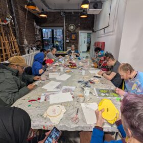 A group of people sat around a table in the OT creative space studio working on creative projects including embroidery and gem art.