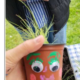 A hand is holding a plant pot which has been painted with a face and goggly eyes. The pot has grass growing out of it.