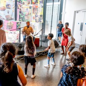 A grups of musicians play their instruments on a small stage as children dance around them.