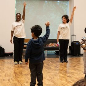 A group of young people in a dance session. They stand with one arm raised up.
