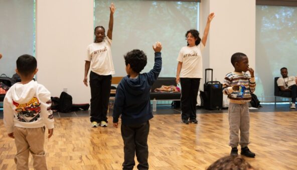 A group of young people in a dance session. They stand with one arm raised up.