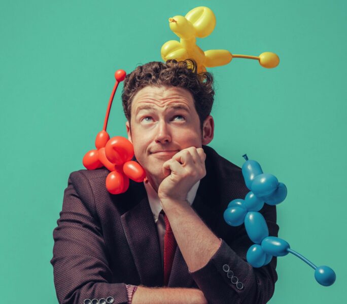 A man with a yellow balloon model on his head, a read balloon model on his shoulder and a blue balloon model on his arm.