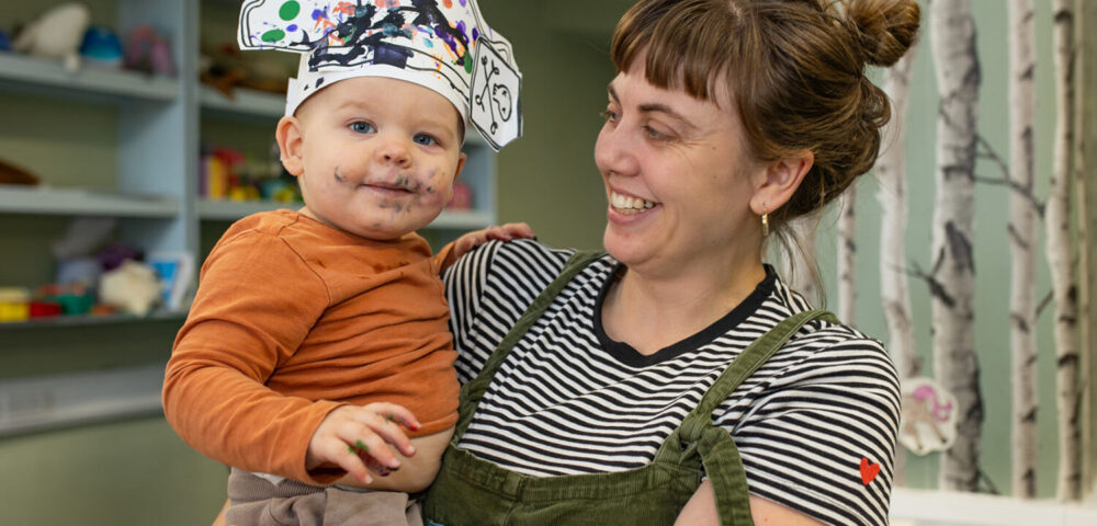 A mother wearing a black and white stripy top holds a baby, who is wearing an orange top and a pirate hat they have painted. The baby also has green paint on its face.