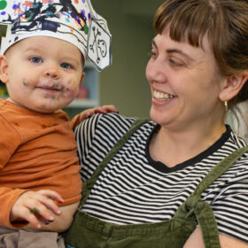 A mother wearing a black and white stripy top holds a baby, who is wearing an orange top and a pirate hat they have painted. The baby also has green paint on its face.