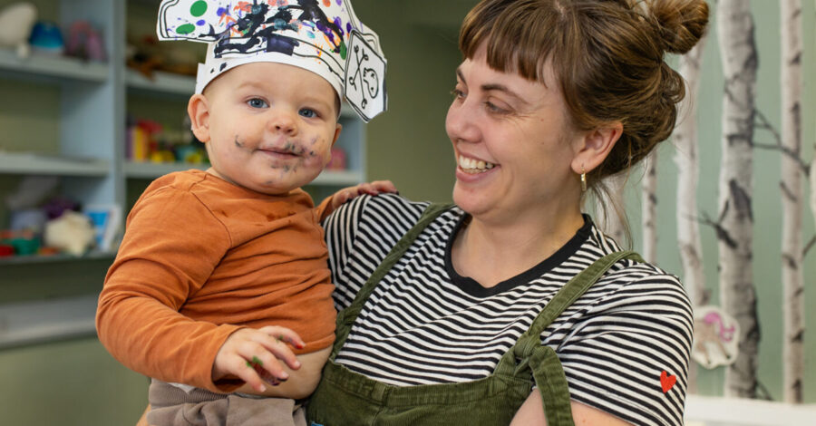 A mother wearing a black and white stripy top holds a baby, who is wearing an orange top and a pirate hat they have painted. The baby also has green paint on its face.