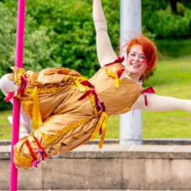 A performer with long red hair, wearing a yellow dress, holds her arms out as she swings from a rope which is wound around her legs.