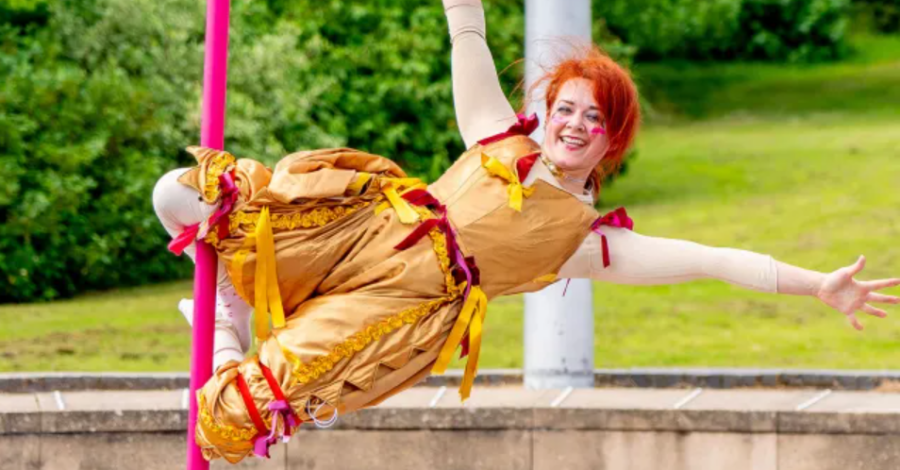 A performer with long red hair, wearing a yellow dress, holds her arms out as she swings from a rope which is wound around her legs.