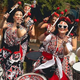 A group of batala drummers in tradition Brazilian Batala costume playing their drums as they walk outside.