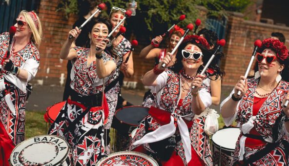 A group of batala drummers in tradition Brazilian Batala costume playing their drums as they walk outside.
